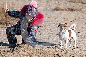 Little girl in hat and jacket in the winter plays on the beach on the sand with a Jack Russell