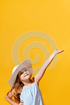 Little girl in a hat and a sundress on a yellow background. The child raised his hand up and points to the copy space