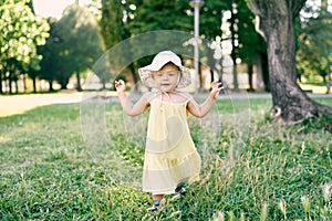 Little girl in a hat walks on the lawn with her hands up