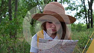 Little girl in a hat and with yellow backpack sits in summer camp in the forest near the tent. Child seriously looking