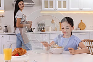 Little girl having breakfast while mother cooking food in kitchen. Single parenting