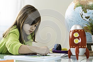 Little girl having fun drawing with wax colors on school notebook for homework - On the desk there are a globe and a red rocket