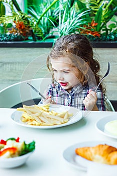 Little girl having lunch in the restaurant with the table knife and fork in hands.