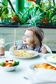 Little girl having lunch in the restaurant with the table knife and fork in hands.