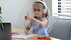 Little girl with headphones shows thumb up to laptop camera