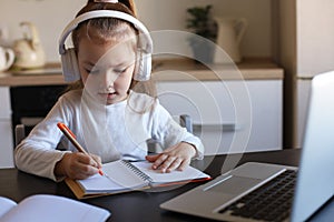 Little girl in headphones sit at desk writing in notebook studying online do exercises at home photo