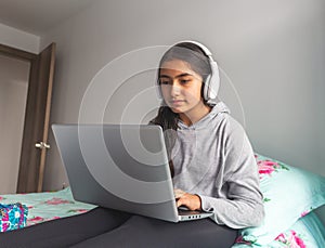 Little girl with headphones sitting on her bed typing on her laptop