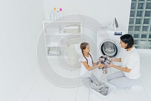 Little girl helper and her mother pose in laundry room near washing machine, sit on white floor, hold dirty shirt, do washing at