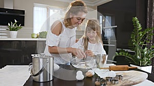 Little girl helping her mom in the kitchen by stirring the ingredients for their cake with a spoon. Little cute girl