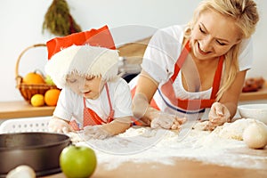 Little girl and her blonde mom in red aprons playing and laughing while kneading the dough in kitchen. Homemade pastry