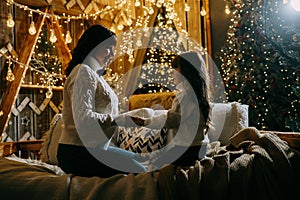 A little girl with her mother in a cozy home environment on the sofa next to the Christmas tree. The theme of New Year photo