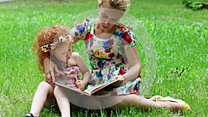 Little girl and her mother read book on grass in summer park