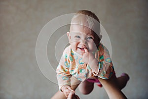 A little girl in her mother`s arms above her head smiles with a finger in her mouth