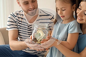 Little girl with her parents holding glass jar with dollars indoors. Money savings concept