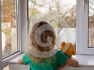 A little girl with her teddy bear looks out the window at the garden.