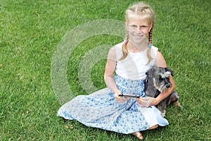 Little girl with his dog sitting on green grass and read e-book.