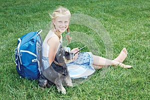 Little girl with his dog sitting on green grass and read e-book.