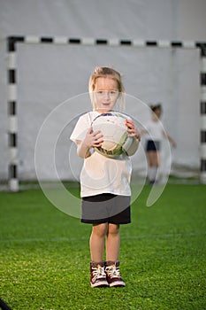 A little girl holding the ball with both hands on the football field, laughing and looking at the camera