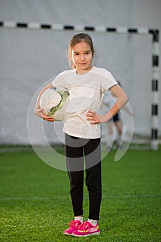 A little girl holding the ball in her hands on the football field, smiling and looking at the camera
