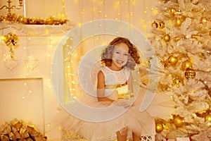 Little Girl Holding Candle sitting next to Christmas Tree Decorated with Golden Ornaments and Lights. Happy Child in White Room