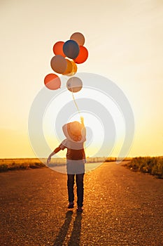 Little girl holding colorful balloons in hand goes into the sunset