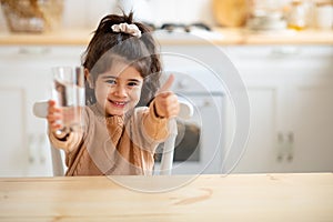 Little Girl Holding Glass Of Water In Kitchen And Showing Thumb Up