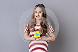 Little girl holding little paper house looking at camera with smile dreaming about apartment.