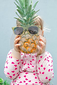 Little girl holding a pineapple with glasses in front of her face. Pineapple instead of the head. In the garden outdoors.