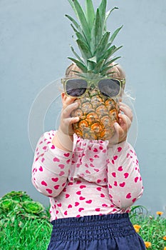 Little girl holding a pineapple with glasses in front of her face. Pineapple instead of the head. In the garden outdoors.