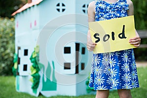 Little Girl Holding Sold Sign Outside Cardboard Playhouse