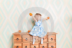 Little girl holding tangerines or oranges in her hands while sitting on the wooden wardrobe. Wearing light blue dress