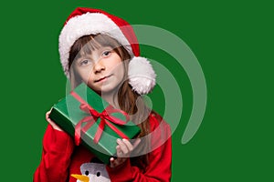 A little girl holds a gift on a green isolated background. A child in a Santa Claus hat and a warm sweater holds a gift