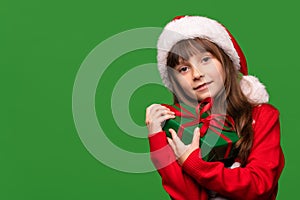 A little girl holds a gift on a green isolated background. A child in a Santa Claus hat and a warm sweater holds a gift