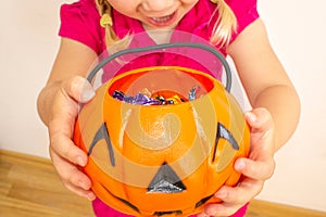 A little girl holds a pumpkin with candy in her hands and stretches it to get even more candies for Halloween. Close-up.