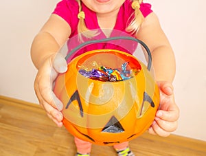 A little girl holds a pumpkin with candy in her hands and stretches it to get even more candies for Halloween. Close-up.