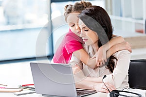 Little girl hugging busy mother working with laptop in office