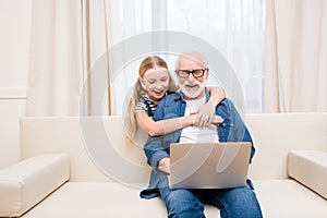 Little girl hugging happy grandfather using laptop at home