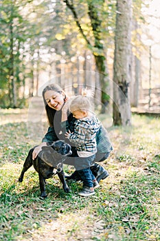 Little girl hugging laughing mom stroking dog in park