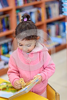 Little Girl Indoors In Front Of Books. Cute Young Toddler Play With Toy and Reading Book.Library, Shop, Shelving In Home