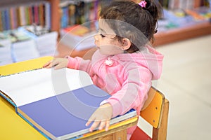 Little Girl Indoors In Front Of Books. Cute Young Toddler Sitting On A Chair Near Table and Reading Book. Library, Shop.