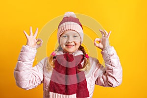 Little girl in a jacket, scarf and hat on a yellow background. The child shows an OK sign with two hands..