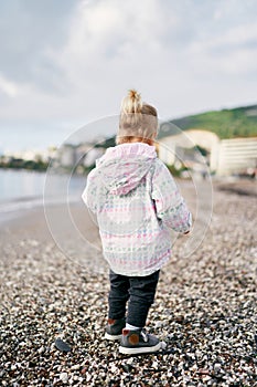 Little girl in a jacket walks along a pebbly beach, looking down at her feet. Back view