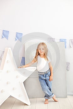 Little girl in jeans and a white T-shirt stands by the luminous star of the lamp. Large decorative retro star. Christmas, New Year