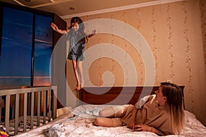 Little girl jumps from the head of the bed, young woman lies, watching her daughter perform