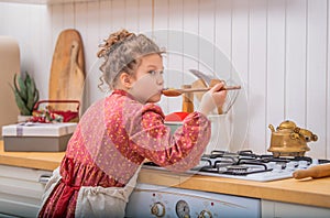 A little girl in the kitchen in an apron cooks something in a saucepan on a gas stove