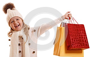 A little girl in a knitted hat with a pompon, scarf and sweater, is holding multicolored paper bags. Isolated on white background