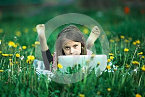 Little girl with laptop lying on green grass.