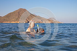 Little girl laughing with diving glasses on back of woman snorkeling in the water of a beach in Andalusia