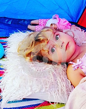 A little girl laying down in a tent resting her head on a fluffy pillow, whilst looking on quite solemnly.