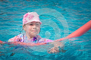 Little girl learns to swim with the help of noodles. The child is trying to swim in the pool. Beautiful baby bathes in turquoise